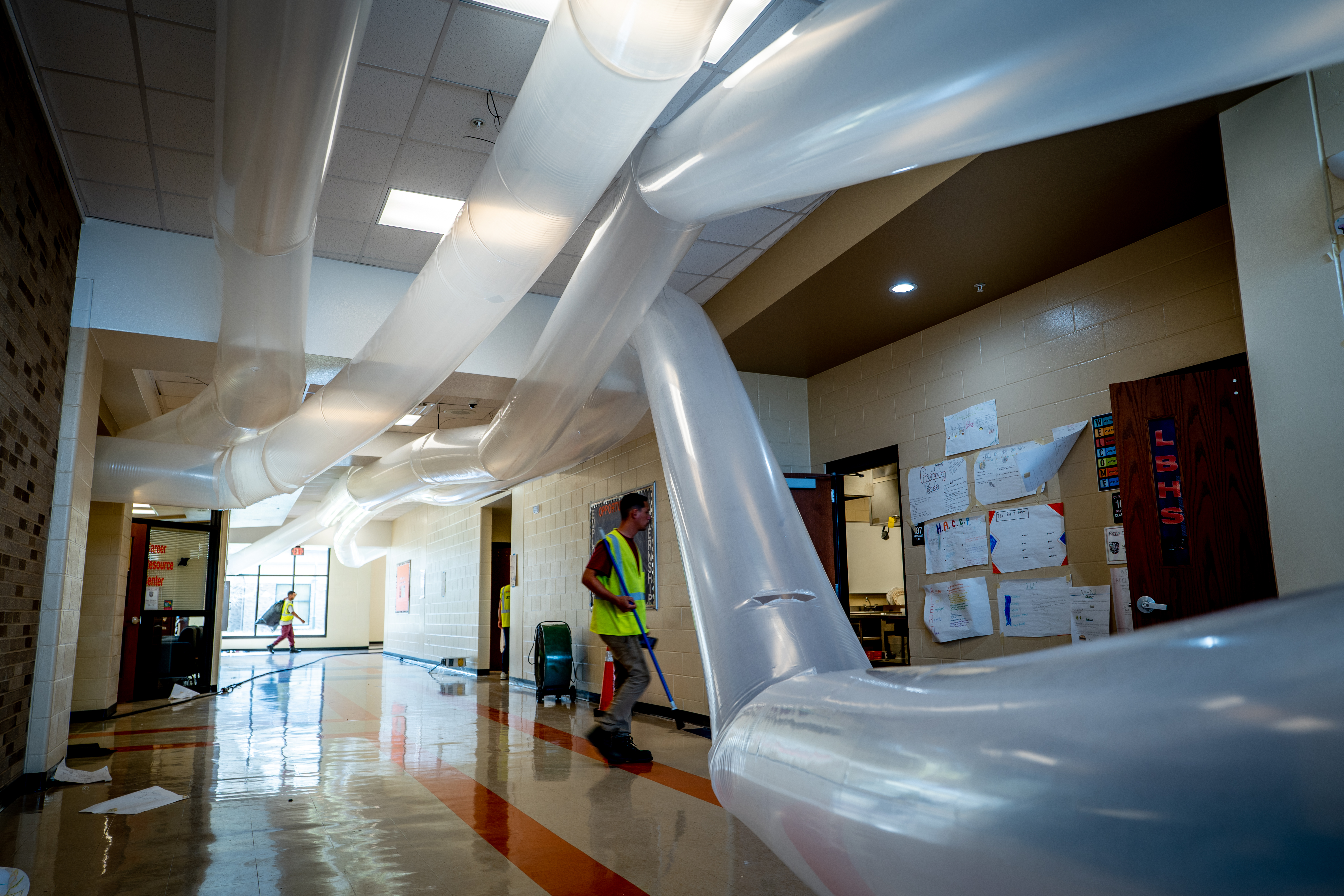 A school hallway with transparent air ducts running along the ceiling and walls. Workers are in the hall, wearing safety vests.