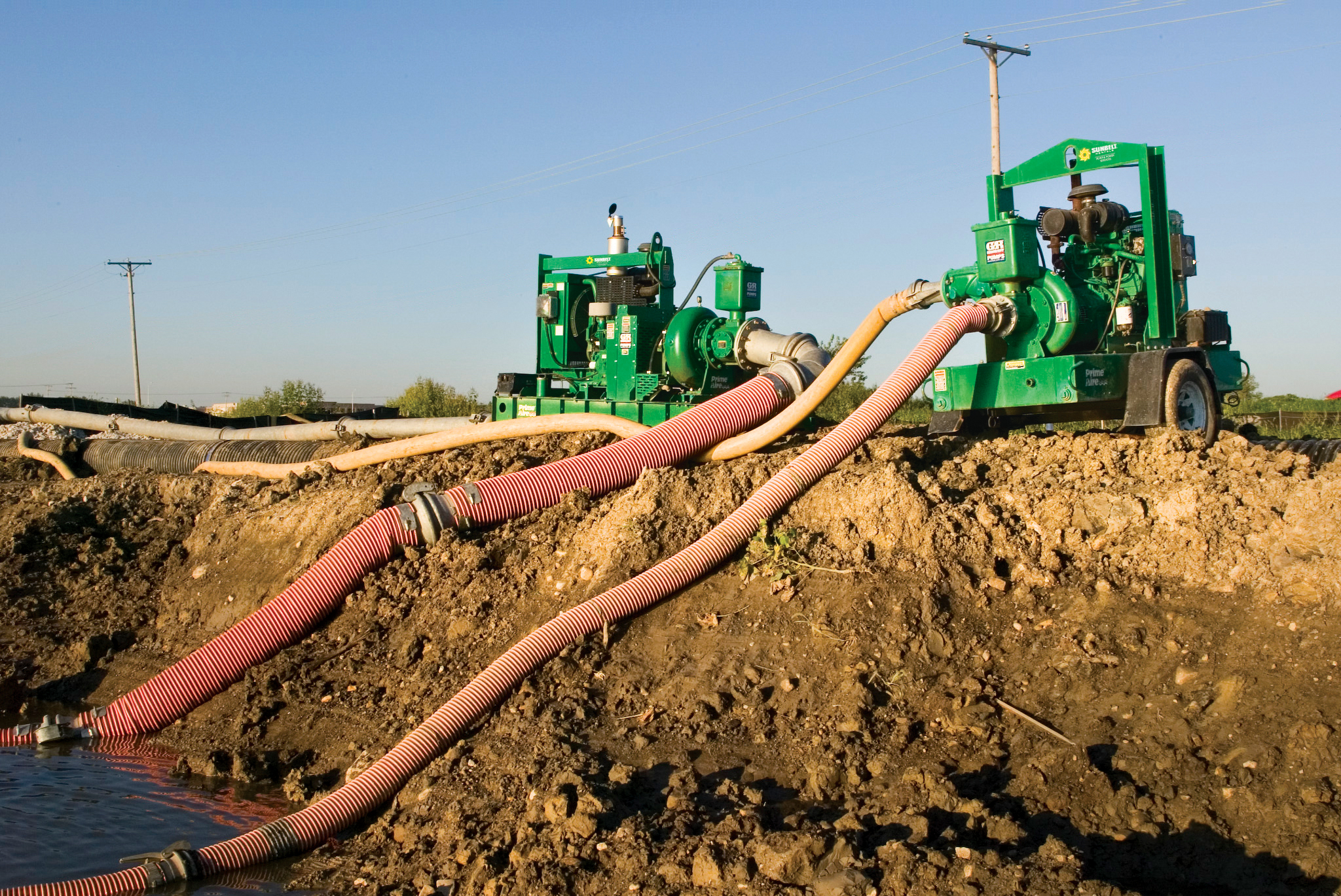 Water pumps from Sunbelt Rentals on a dirt embankment, connected by hoses to a nearby water source.