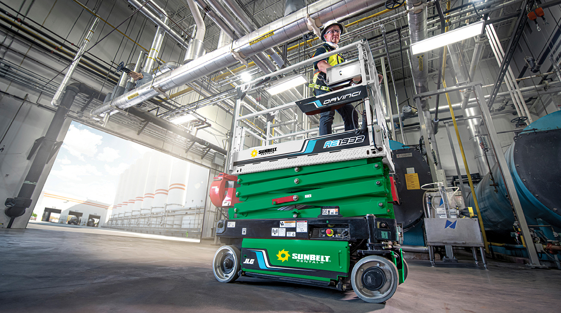 Someone standing on a collapsed scissor lift, inside of a large and open building.