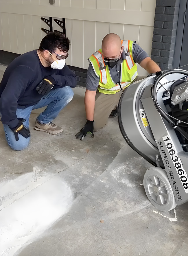 Two people wearing safety equipment, with a concrete grinder in a garage.