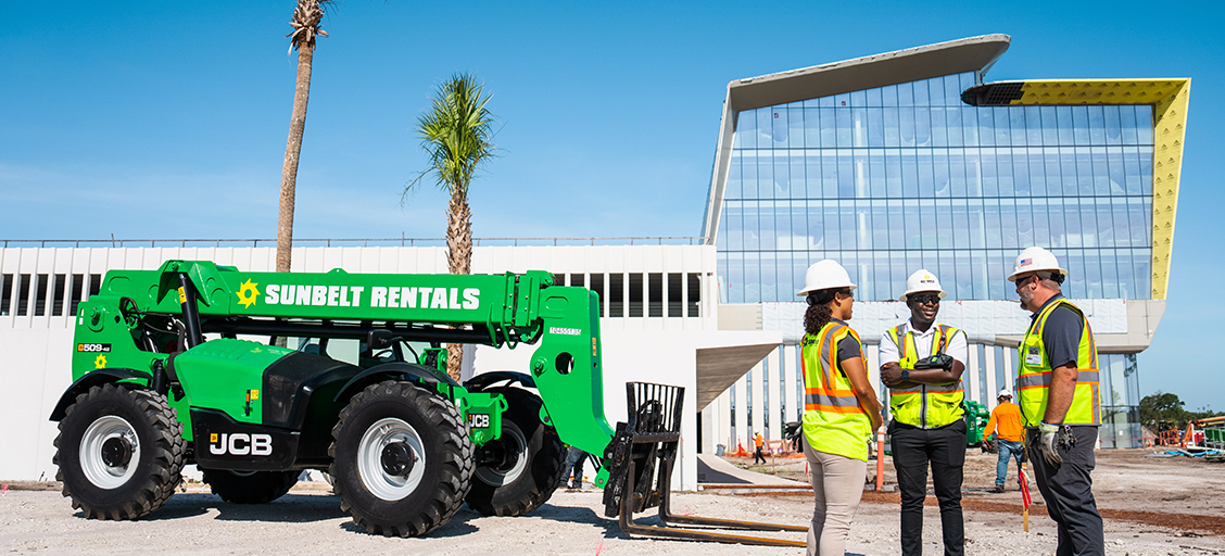 Three builders wearing fluorescent vests, next to a Sunbelt Rentals telehandler, outside of a warehouse facility.