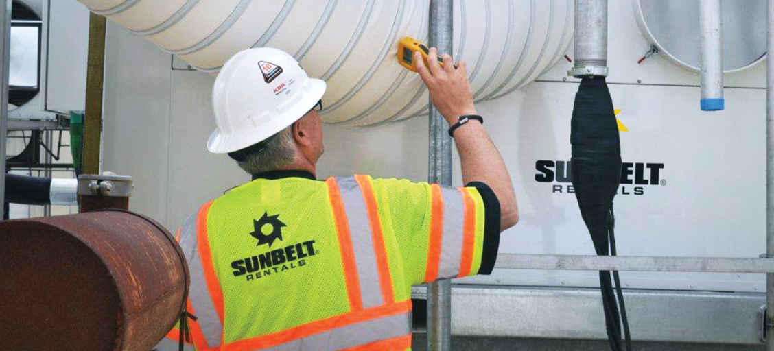 A person wearing a white construction helmet and Sunbelt Rentals safety vest uses a portable measurement tool to monitor the air temperature in ducting connected to a portable air conditioner.