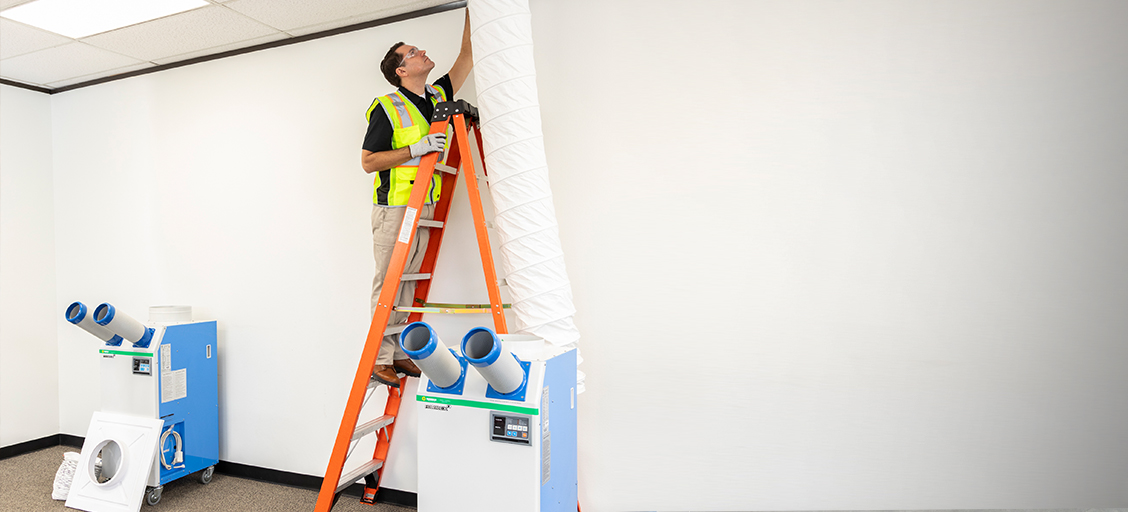A man in a safety vest stands on an orange ladder to secure white ducting that connects a portable air conditioner to a drop ceiling.